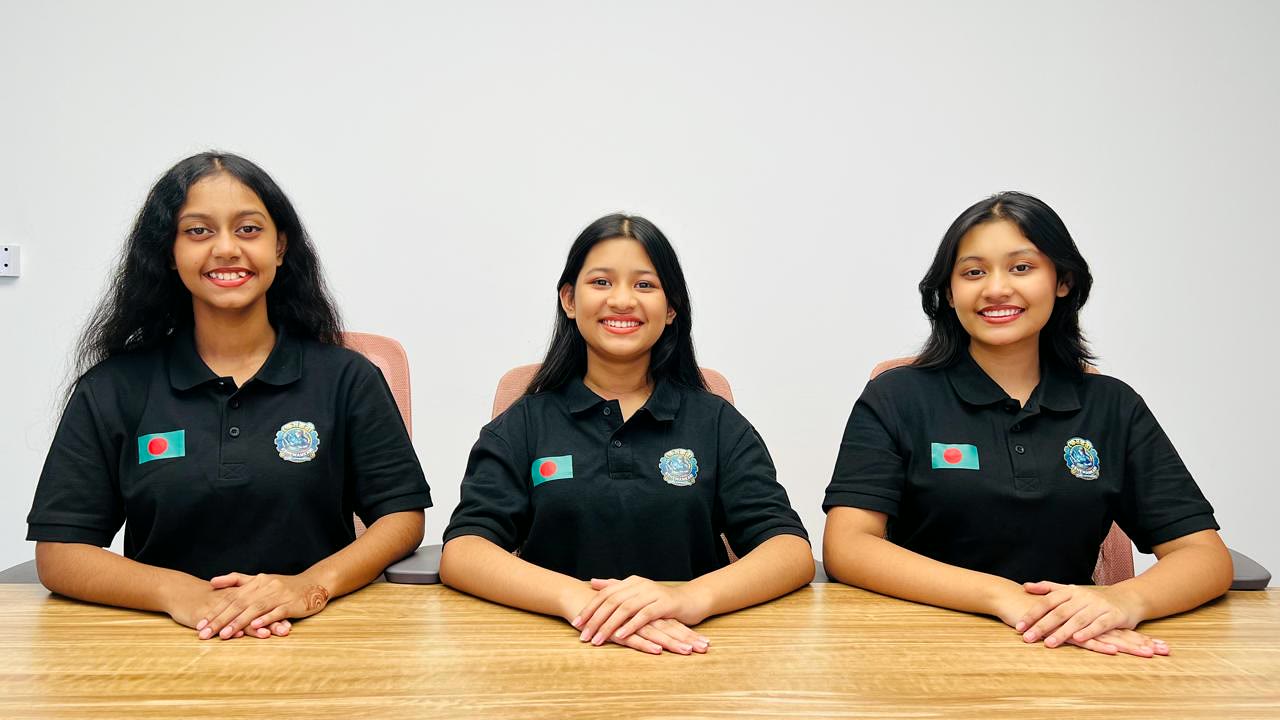 The three members of Team Kshemankari seated at a table, in black team polo shirts carrying the Bangladesh flag and their school crest.